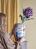 A woman is holding a decorative vase with a flower in it, in front of a yellow curtain. Náhled obrázku produktu