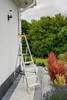 A ladder is placed next to a white house wall, with a garden in the background. Náhľad obrázku produktu