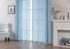 A room interior with a large window covered by light blue curtains, a white armchair, and a white fireplace with books and a lamp. Produktbild-Vorschau