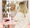 A woman is applying makeup in front of a vanity mirror with lights. Voir les images du produit