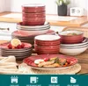 A collection of red and white striped ceramic dishes and bowls are arranged on a wooden table, with some food items and a lemon slice visible on one of the plates. The background shows a kitchen counter and a window. Náhled obrázku produktu