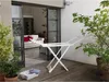 A white Vileda Solar drying rack is set up on a wooden deck outside a home, with plants and an open doorway to an interior living space visible. Náhled obrázku produktu