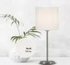 A white table lamp with a fabric shade is displayed on a white surface in front of a textured light gray background. A decorative white vase with a plant is to the left of the lamp. A circular inset shows a close-up of the white material with text. Produktbild-Vorschau