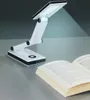 A white LED desk lamp is positioned on a gray surface next to an open book. Produktbild-Vorschau