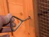 A hand is shown holding a metal latch to a wooden structure, possibly a chicken coop or animal enclosure. Produktbild-Vorschau