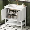 A white bathroom vanity with open doors and shelves, displaying various toiletries and cleaning products. A gold faucet is visible on the white sink. Náhľad obrázku produktu