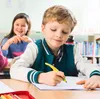 Two children are in a classroom setting, with one boy in the foreground writing in a notebook and a girl in the background smiling. Anteprima dell’immagine prodotto