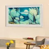 A Samsung Frame TV displaying a painting of a landscape with trees and a moon, mounted on a cream-colored wall above a small table with books and a vase of yellow flowers. Produktbild-Vorschau