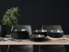 A table setting with a stack of plates and bowls, a vase with greenery, and chairs in the background. Produktbild-Vorschau