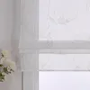 A close-up view of a white sheer roman blind with a delicate gold floral pattern, partially covering a window. A few white flowers are visible on the left side of the image. Produktbild-Vorschau