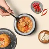 A top-down view of a meal being prepared and served, with a hand holding a fork to scoop rice from a small pot, surrounded by ingredients like chilies and cheese. Produktbild-Vorschau