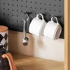A close-up view of a pegboard with hooks holding two white mugs and a metal scoop. A patterned canister and part of a kettle are visible on the shelf below. Produktbild-Vorschau