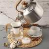A kettle pouring hot water into a teapot, with a teacup and cookies nearby. Voir les images du produit