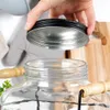 A hand is shown holding a black lid above a glass beverage dispenser. Produktbild-Vorschau