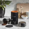 A coffee grinder and its parts are displayed on a kitchen counter with a plant and other kitchen items in the background. Náhled obrázku produktu