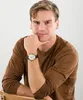A man wearing a brown long-sleeved shirt and a watch is posing for a photo against a white background. Produktbild-Vorschau