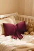 A burgundy bow-shaped pillow and a teddy bear are placed on a bed with white bedding. Produktbild-Vorschau