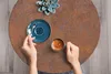 A top-down view of a person holding a cup of coffee and a spoon on a round, rusty-looking table. Náhľad obrázku produktu