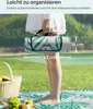 A person holding a rolled-up picnic blanket with tropical leaf pattern, with a picnic basket and food in the foreground. Produktbild-Vorschau