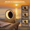 A wooden sunrise alarm clock is placed on a woven table next to a book, with a beach and sunset in the background. Several inset images show the alarm clock at different brightness levels. Produktbild-Vorschau