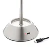 A close-up of the base of a silver LED table lamp, showing a USB-C port and a red indicator light, with a USB-C cable connector in the foreground. Produktbild-Vorschau