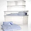 A stack of clear plastic storage boxes with a blue shirt folded in the bottom box and another blue shirt folded inside one of the upper boxes. Produktbild-Vorschau