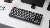 A black mechanical keyboard with gray and orange keycaps is placed on a white desk, with a pair of glasses and a black dial in the background. Produktbild-Vorschau