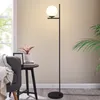 A modern floor lamp with a white globe shade stands next to a chair and a small side table with a plant and picture frame. Anteprima dell’immagine prodotto