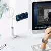 A white flexible phone holder is clamped to a desk next to a computer monitor. A person's hand is visible on the right, typing on a keyboard. Podgląd zdjęcia produktu