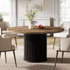 A round dining table with a wooden top and a black fluted base, surrounded by chairs in a dining room setting. Produktbild-Vorschau