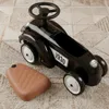 A black retro ride-on car for children is shown on a carpeted floor, with a brown padded seat accessory next to it. Náhled obrázku produktu
