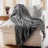 A gray blanket is draped over a white armchair with a textured pillow. A small wooden table with a potted plant is to the left. Produktbild-Vorschau