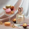 A perfume bottle is displayed with macarons and chocolates, suggesting a sweet and luxurious theme. Voir les images du produit