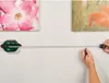 A person is using a digital laser level with a tape measure to mark a point on a wall between two pictures. Produktbild-Vorschau