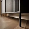 Close-up of the bottom part of a black cabinet with vertical slats, resting on thin black legs on a wooden floor. Produktbild-Vorschau