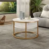 A round coffee table with a marble-like top and gold frame is placed on a gray rug in a living room setting. Sunlight streams in from a large window, illuminating a plant and a sofa in the background. Produktbild-Vorschau