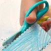 A child's hand is using teal-colored training scissors to cut along a dotted line on a piece of paper that has been colored with blue crayon. Voir les images du produit