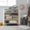 A white cabinet with shelves filled with folded clothes and books, placed in a room with a chair and a hat on the wall. Náhled obrázku produktu