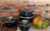 A set of black pots and pans with glass lids, along with a pumpkin, are displayed on a wooden surface in front of a brick wall. Produktbild-Vorschau