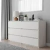 A white dresser with six drawers is shown in a bedroom setting, with a lamp, picture frames, and books on top. A grey curtain and rug are also visible. Náhled obrázku produktu