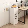 A white filing cabinet with three drawers is placed next to a wooden desk in a home office setting. The cabinet has black handles. On top of the cabinet are several books and a desk calendar. The wall behind the cabinet has some papers taped to it. Náhled obrázku produktu