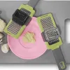 Two garlic graters are shown on a gray surface with some garlic cloves and a pink plate with grated garlic. Produktbild-Vorschau