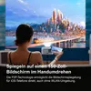 A woman is sitting on a couch, holding a smartphone and looking at a large projected image on a screen. A mini projector is on a tripod in front of her. Produktbild-Vorschau