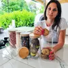 Klare Glas-Vorratsgläser mit Bambusdeckeln auf einer weißen Arbeitsplatte, gefüllt mit verschiedenen Lebensmitteln, eine Frau hält einen Keks Produktbild-Vorschau