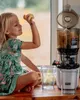 A young girl is holding a piece of fruit and looking at a juicer on a kitchen counter. Produktbild-Vorschau