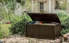 A brown storage box with its lid open, filled with garden tools, sits in a lush garden setting next to a house. Podgląd zdjęcia produktu