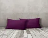 Two purple pillows are placed on a wooden floor in front of a textured gray wall. Produktbild-Vorschau