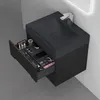 A black vanity cabinet with an open drawer displaying makeup and jewelry, mounted on a white wall with a gray tiled floor. Produktbild-Vorschau