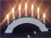 A white arched candle holder with seven electric candles and green pine sprigs, displayed on a dark surface. Voir les images du produit