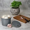 A grey ceramic garlic pot with a lid, filled with garlic bulbs, sits on a light grey surface next to a wooden cutting board with chopped garlic and a knife. A potted basil plant in a woven basket is in the background. Produktbild-Vorschau
